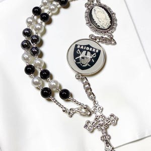 May include: A rosary with black, white, and gray beads, a silver cross, and two charms. One charm features a cameo of a woman, and the other displays the Raiders logo in black and white.