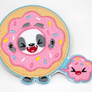 以下が含まれることがあります： ピンクのドーナツの中にパンダが笑顔で描かれた刺繍ワッペン。小さなカップケーキのキャラクターが吹き出し付きで付いています。ワッペンは水色の縁取りで、白い背景にあります。