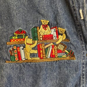 Peut inclure: Chemise en jean brod&eacute;e avec un motif fantaisiste d'ours en peluche sur des &eacute;tag&egrave;res. Les ours portent des n&oelig;uds rouges et sont entour&eacute;s de livres color&eacute;s. La broderie est d&eacute;taill&eacute;e et ajoute une touche ludique au tissu en jean.