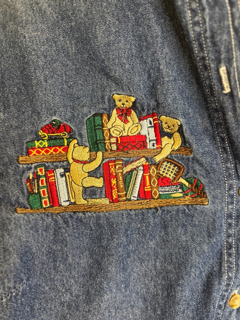 Peut inclure: Chemise en jean brod&eacute;e avec un motif fantaisiste d'ours en peluche sur des &eacute;tag&egrave;res. Les ours portent des n&oelig;uds rouges et sont entour&eacute;s de livres color&eacute;s. La broderie est d&eacute;taill&eacute;e et ajoute une touche ludique au tissu en jean.
