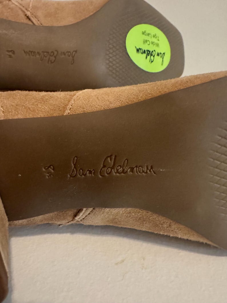K&ouml;nnte beinhalten: Nahaufnahme der Sohle eines Paares hellbrauner Wildlederstiefel. Der Markenname "Sam Edelman" ist auf die Sohle gepr&auml;gt. Ein gr&uuml;ner Aufkleber zeigt "Weiter Waden, Gr&ouml;&szlig;e Large".