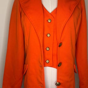 Puede incluir: Conjunto vintage de blazer y chaleco de color naranja brillante. El blazer tiene solapas clásicas y dos bolsillos delanteros. El chaleco tiene botones dorados. El conjunto se exhibe en un maniquí.