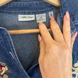 Peut inclure: Une chemise en jean bleu avec des d&eacute;tails brod&eacute;s. La chemise pr&eacute;sente un devant boutonn&eacute;, un col et une &eacute;tiquette qui indique "CABINCREEK". La chemise est suspendue &agrave; un cintre en bois.