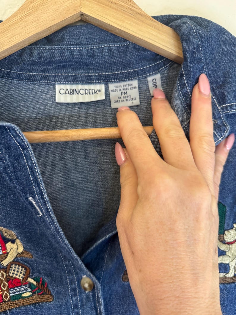 Peut inclure: Une chemise en jean bleu avec des d&eacute;tails brod&eacute;s. La chemise pr&eacute;sente un devant boutonn&eacute;, un col et une &eacute;tiquette qui indique "CABINCREEK". La chemise est suspendue &agrave; un cintre en bois.