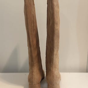 K&ouml;nnte beinhalten: Ein Paar kniehohe Stiefel aus hellbraunem Wildleder. Die Stiefel haben einen Blockabsatz und einen goldenen Rei&szlig;verschluss. Sie stehen auf einer wei&szlig;en Oberfl&auml;che.