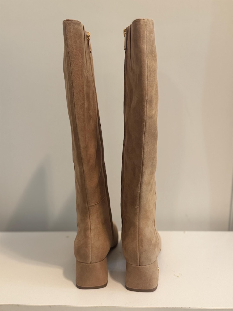 K&ouml;nnte beinhalten: Ein Paar kniehohe Stiefel aus hellbraunem Wildleder. Die Stiefel haben einen Blockabsatz und einen goldenen Rei&szlig;verschluss. Sie stehen auf einer wei&szlig;en Oberfl&auml;che.