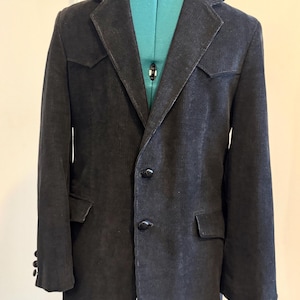 Puede incluir: Una blazer de pana gris oscuro con solapa de muesca, dos bolsillos delanteros y tres botones en cada puño. La blazer tiene cierre de dos botones y se muestra sobre un maniquí turquesa. La tela tiene una textura acanalada vertical.