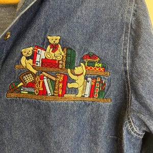 Peut inclure: Chemise en jean bleu avec une broderie de nounours sur une &eacute;tag&egrave;re de livres. Les ours sont dans diverses poses, avec des livres rouges, verts et blancs. La chemise a une fermeture boutonn&eacute;e et un col.