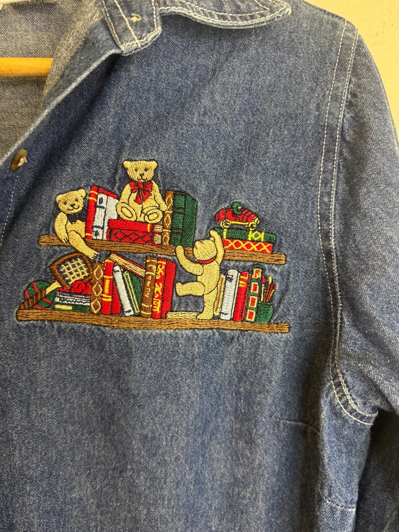 Peut inclure: Chemise en jean bleu avec une broderie de nounours sur une &eacute;tag&egrave;re de livres. Les ours sont dans diverses poses, avec des livres rouges, verts et blancs. La chemise a une fermeture boutonn&eacute;e et un col.