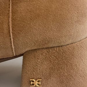 K&ouml;nnte beinhalten: Nahaufnahme eines beigefarbenen Wildlederstiefels mit Blockabsatz. Der Stiefel hat ein goldfarbenes Logo auf der Ferse. Das Wildledermaterial hat ein weiches, strukturiertes Aussehen. Die N&auml;hte sind an der Seite des Stiefels sichtbar.