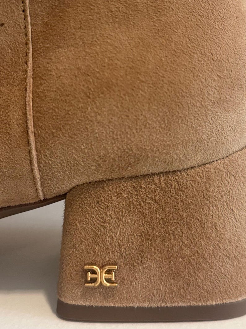 K&ouml;nnte beinhalten: Nahaufnahme eines beigefarbenen Wildlederstiefels mit Blockabsatz. Der Stiefel hat ein goldfarbenes Logo auf der Ferse. Das Wildledermaterial hat ein weiches, strukturiertes Aussehen. Die N&auml;hte sind an der Seite des Stiefels sichtbar.
