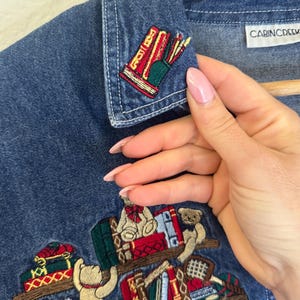 Peut inclure: Chemise en jean bleu avec des broderies de biblioth&egrave;ques, de livres et d'ours en peluche. La chemise a un col et le nom de la marque "CABINCREEK" sur l'&eacute;tiquette. La broderie comprend des accents rouges, verts et dor&eacute;s.