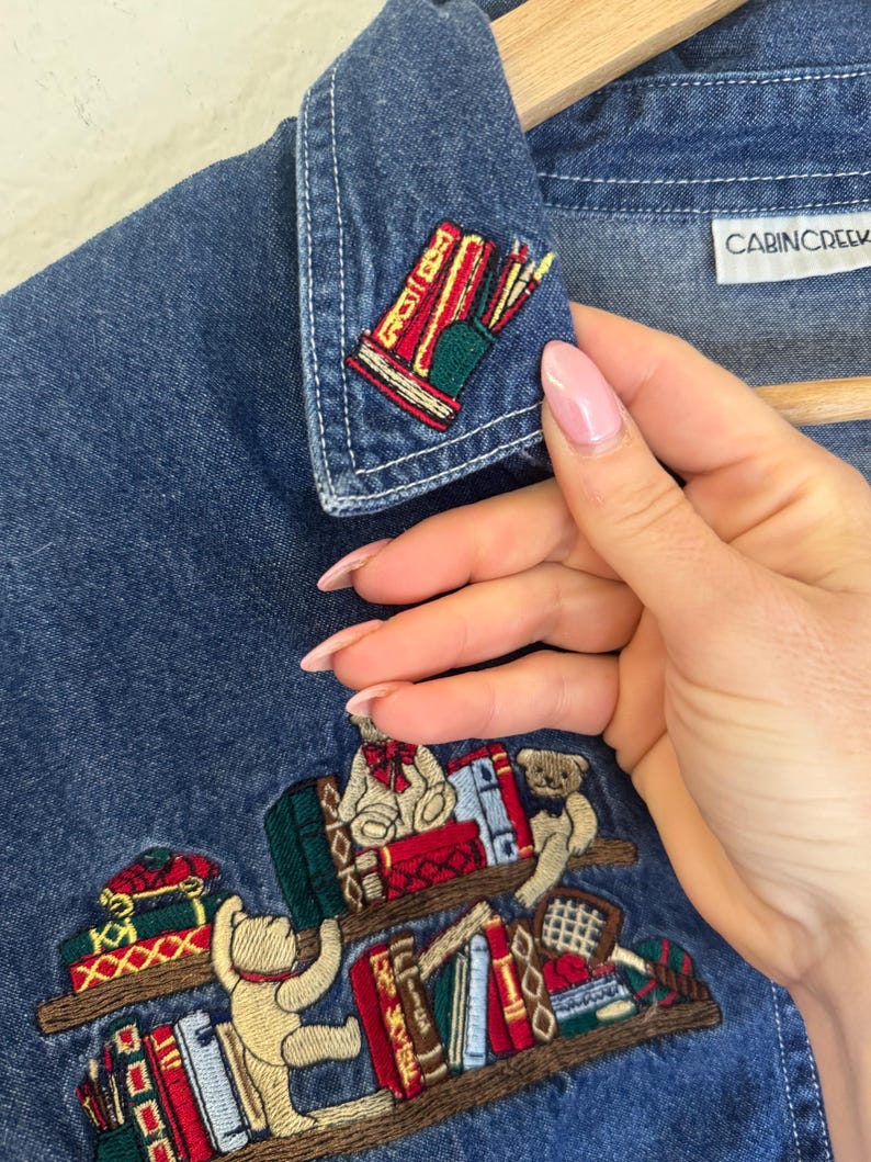 Peut inclure: Chemise en jean bleu avec des broderies de biblioth&egrave;ques, de livres et d'ours en peluche. La chemise a un col et le nom de la marque "CABINCREEK" sur l'&eacute;tiquette. La broderie comprend des accents rouges, verts et dor&eacute;s.