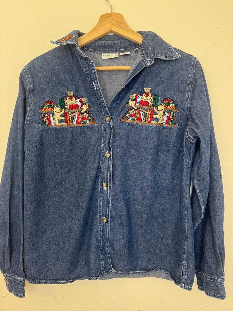 Peut inclure: Chemise en jean bleu avec des manches longues et un col. La chemise est orn&eacute;e de broderies de petits ours lisant des livres sur la poitrine. Les broderies incluent des livres rouges, verts et jaunes. Boutons en bois.
