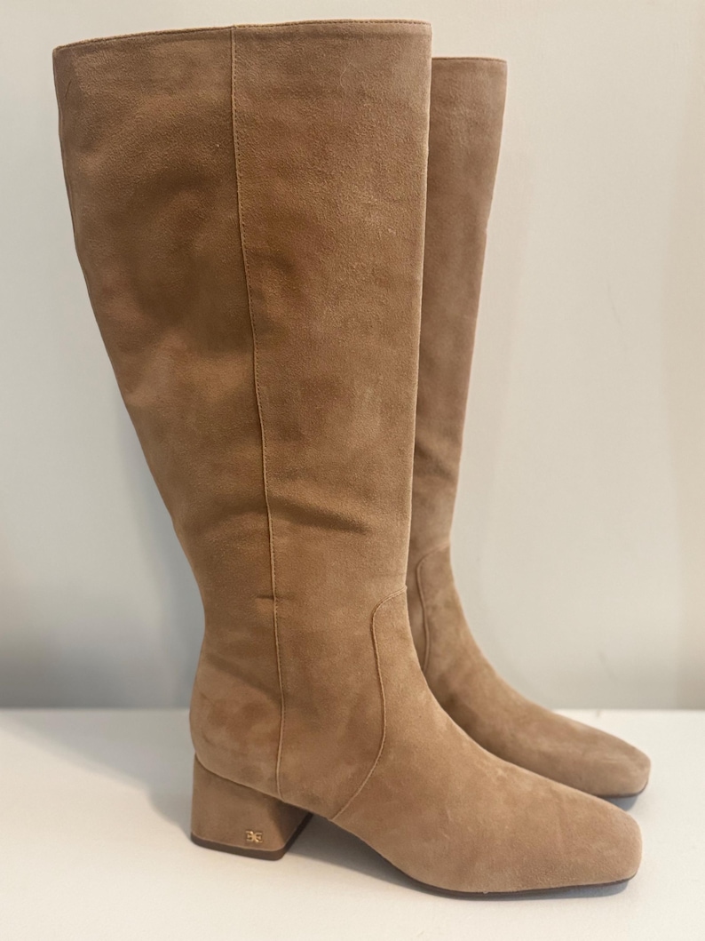 K&ouml;nnte beinhalten: Ein Paar kniehohe Stiefel aus beigefarbenem Wildleder. Die Stiefel haben eine quadratische Spitze, einen Blockabsatz und ein dezentes Logo auf der Ferse. Die Stiefel sind aus weichem, strukturiertem Material.