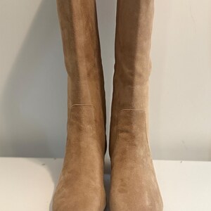 K&ouml;nnte beinhalten: Ein Paar kniehohe Stiefel aus beigefarbenem Wildleder. Die Stiefel haben eine quadratische Spitze und einen Blockabsatz. Die Stiefel sind aus weichem Wildledermaterial und haben eine neutrale Farbe. Die Stiefel stehen auf einer wei&szlig;en Oberfl&auml;che.