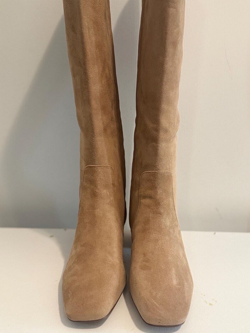K&ouml;nnte beinhalten: Ein Paar kniehohe Stiefel aus beigefarbenem Wildleder. Die Stiefel haben eine quadratische Spitze und einen Blockabsatz. Die Stiefel sind aus weichem Wildledermaterial und haben eine neutrale Farbe. Die Stiefel stehen auf einer wei&szlig;en Oberfl&auml;che.