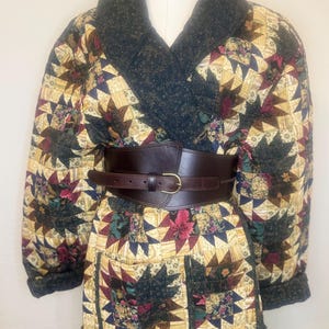 Puede incluir: Chaqueta acolchada con un estampado de estrella en tonos dorados, verdes y burdeos. La chaqueta tiene cuello y puños verde oscuro, y un cinturón de cuero marrón ciñe la cintura. La chaqueta presenta dos bolsillos y un diseño único.