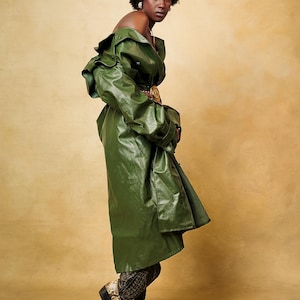 May include: An olive green trench coat with a glossy finish, worn off the shoulders, paired with snakeskin-patterned cowboy boots featuring black leather detailing and a low heel. The coat has wide lapels and long sleeves, creating a layered look.