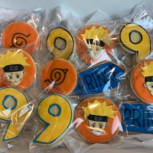 May include: A box of decorated cookies featuring cartoon character faces, orange swirls, and the number 9. Some cookies have the word "PRINCE" or "RING" on them. The cookies are individually wrapped.