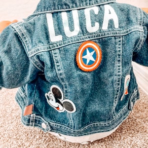 May include: A denim jacket with the name "LUCA" in white block letters on the back. The jacket features patches of Mickey Mouse and Captain America's shield. The jacket is a medium wash blue.