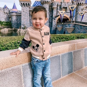 May include: A tan and black bomber jacket with a Mickey Mouse patch and the name "LEO" on the chest. The jacket is paired with blue jeans and black and white sneakers. The background features a castle.