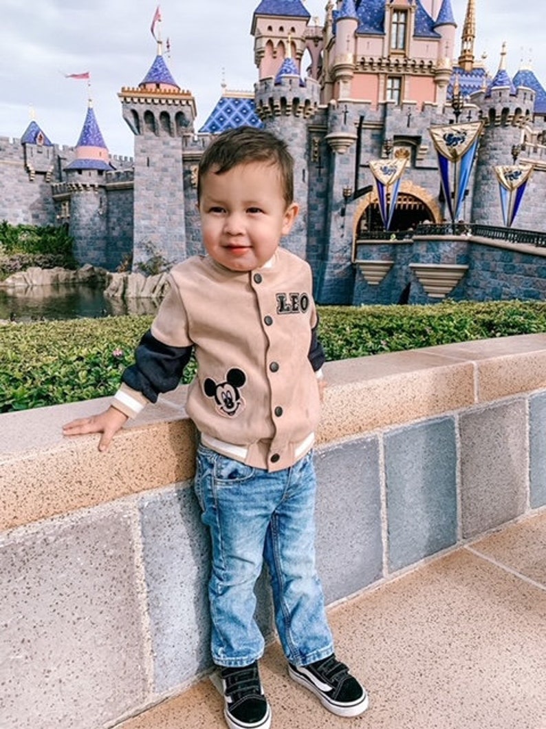 May include: A tan and black bomber jacket with a Mickey Mouse patch and the name "LEO" on the chest. The jacket is paired with blue jeans and black and white sneakers. The background features a castle.