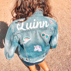 May include: A light blue denim jacket with the name "Quinn" in white script across the back. The jacket is decorated with patches including a rainbow, sunglasses, and a unicorn. The jacket is worn by a child.