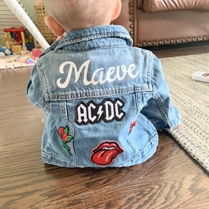 May include: A light blue denim jacket with the name "Maeve" in white script across the back. Patches include "AC/DC", a red rose, and a red tongue graphic. The jacket is designed for a baby or toddler.