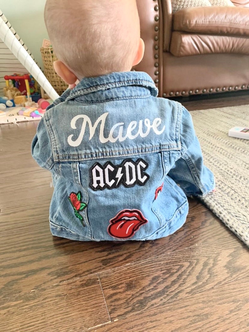 May include: A light blue denim jacket with the name "Maeve" in white script across the back. Patches include "AC/DC", a red rose, and a red tongue graphic. The jacket is designed for a baby or toddler.