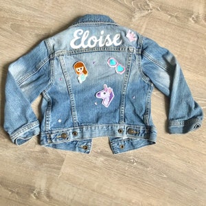 May include: A light blue denim jacket with the name "Eloise" in white script across the back. The jacket is decorated with patches of a mermaid, sunglasses, a unicorn, and a crown. The jacket has long sleeves and metal button closures.