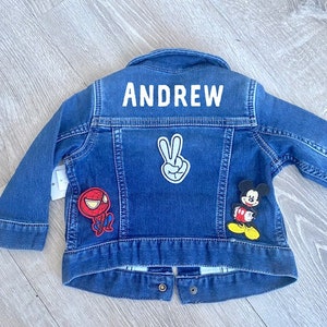 May include: A blue denim jacket with the name "ANDREW" in white block letters across the back. The jacket features patches of Spider-Man, Mickey Mouse, and a peace sign. The jacket has long sleeves and button closures.