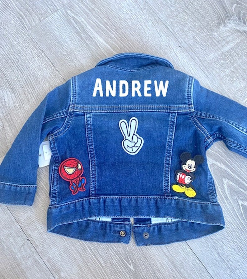 May include: A blue denim jacket with the name "ANDREW" in white block letters across the back. The jacket features patches of Spider-Man, Mickey Mouse, and a peace sign. The jacket has long sleeves and button closures.