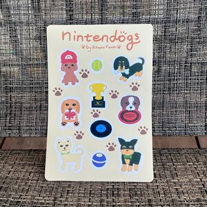 May include: A sticker sheet featuring various cartoon dog illustrations with accessories like a red cap, tennis ball, and trophy. The text "Nintendogs" is at the top, with "by Kitsune Forest" below. Paw prints and a vinyl record are also included.