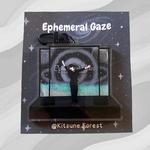 May include: A black acrylic keyring with the text "Ephemeral Gaze" and a silhouette of a person with arms raised. The keyring has a black clasp and is set against a dark background with stars. The design includes the text "@Kitsune Forest".
