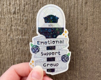 May include: Holographic sticker with the text "Emotional Support Group" in a banner design. The sticker features a dark blue and black design with stars and a rose graphic. The sticker is held by a hand.