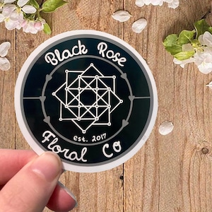 May include: A round sticker with the text "Black Rose Floral Co" in white script. The sticker has a dark teal background with a white geometric design in the center. The text "est. 2017" is also included.