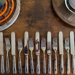 Puede incluir: Un surtido de tenedores y cuchillos plateados dispuestos sobre una superficie de madera. La cubertería tiene detalles ornamentales en los mangos. La imagen también incluye bandejas de madera decorativas y un salvamanteles tejido.
