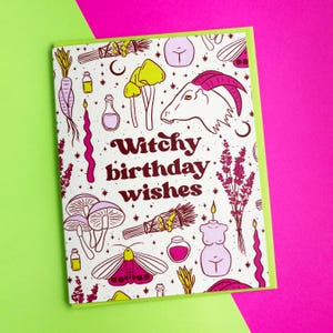 May include: A white card with a pink and yellow illustration of various witchy items, including a goat, mushrooms, candles, and a moth. The text "Witchy birthday wishes" is written in pink letters.