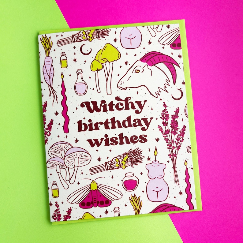 May include: A white card with a pink and yellow illustration of various witchy items, including a goat, mushrooms, candles, and a moth. The text "Witchy birthday wishes" is written in pink letters.