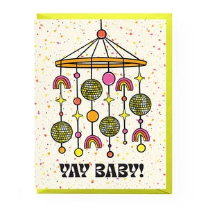 Puede incluir: Una tarjeta de felicitaci&oacute;n con un borde amarillo ne&oacute;n y un fondo color crema salpicado de motas coloridas. La tarjeta presenta un m&oacute;vil de estilo retro con bolas de discoteca, arco&iacute;ris y estrellas. El texto "YAY BABY!" est&aacute; impreso en negrita negra.