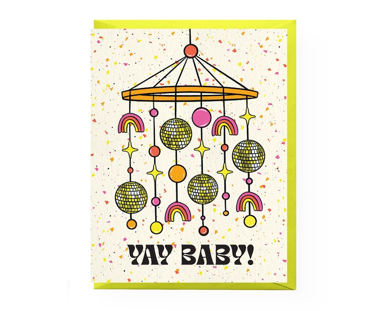 Puede incluir: Una tarjeta de felicitaci&oacute;n con un borde amarillo ne&oacute;n y un fondo color crema salpicado de motas coloridas. La tarjeta presenta un m&oacute;vil de estilo retro con bolas de discoteca, arco&iacute;ris y estrellas. El texto "YAY BABY!" est&aacute; impreso en negrita negra.