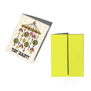 Puede incluir: Tarjeta de felicitaci&oacute;n con un dise&ntilde;o de m&oacute;vil para beb&eacute; y el texto "YAY BABY!". La tarjeta presenta un fondo amarillo con una ilustraci&oacute;n de m&oacute;vil para beb&eacute; en rosa, naranja y amarillo. Una segunda tarjeta est&aacute; abierta, mostrando un interior amarillo brillante.