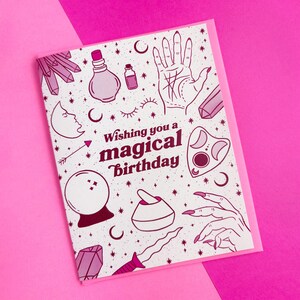 May include: A white card with a pink border features a pink and white illustration of various magical objects, including a crystal, a potion bottle, a hand with lines, a crescent moon, a crystal ball, a mortar and pestle, a dagger, and a hand with long nails. The text "Wishing you a magical birthday" is printed in pink on the card.