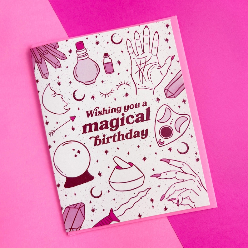 May include: A white card with a pink border features a pink and white illustration of various magical objects, including a crystal, a potion bottle, a hand with lines, a crescent moon, a crystal ball, a mortar and pestle, a dagger, and a hand with long nails. The text "Wishing you a magical birthday" is printed in pink on the card.