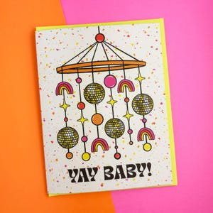 Puede incluir: Tarjeta de felicitaci&oacute;n con borde amarillo ne&oacute;n e ilustraci&oacute;n de m&oacute;vil para beb&eacute;. El m&oacute;vil presenta bolas de discoteca, arco&iacute;ris y estrellas en tonos rosa, naranja y amarillo. Las palabras "YAY BABY!" est&aacute;n impresas en negro en la parte inferior.