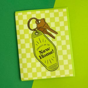 May include: A yellow and green greeting card with a checkered pattern. The card features a cartoon illustration of a motel key with the text "Congrats on the New Home!"