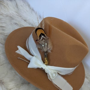Può includere: Un cappello fedora in feltro marrone con un nastro bianco e un accento di piuma. Il cappello è decorato con un piccolo spillo dorato.