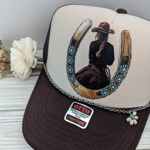 May include: A brown and white trucker hat with a horseshoe and a woman riding a horse design. The hat has a silver chain with a flower charm attached. The hat has a label that says "OTTO WORLD FAMOUS FOAM FRONT OSFM TRUCKER".