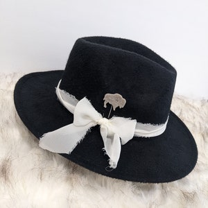 May include: Black fedora hat with a white ribbon and a small buffalo pin. The hat is sitting on a white fluffy surface.